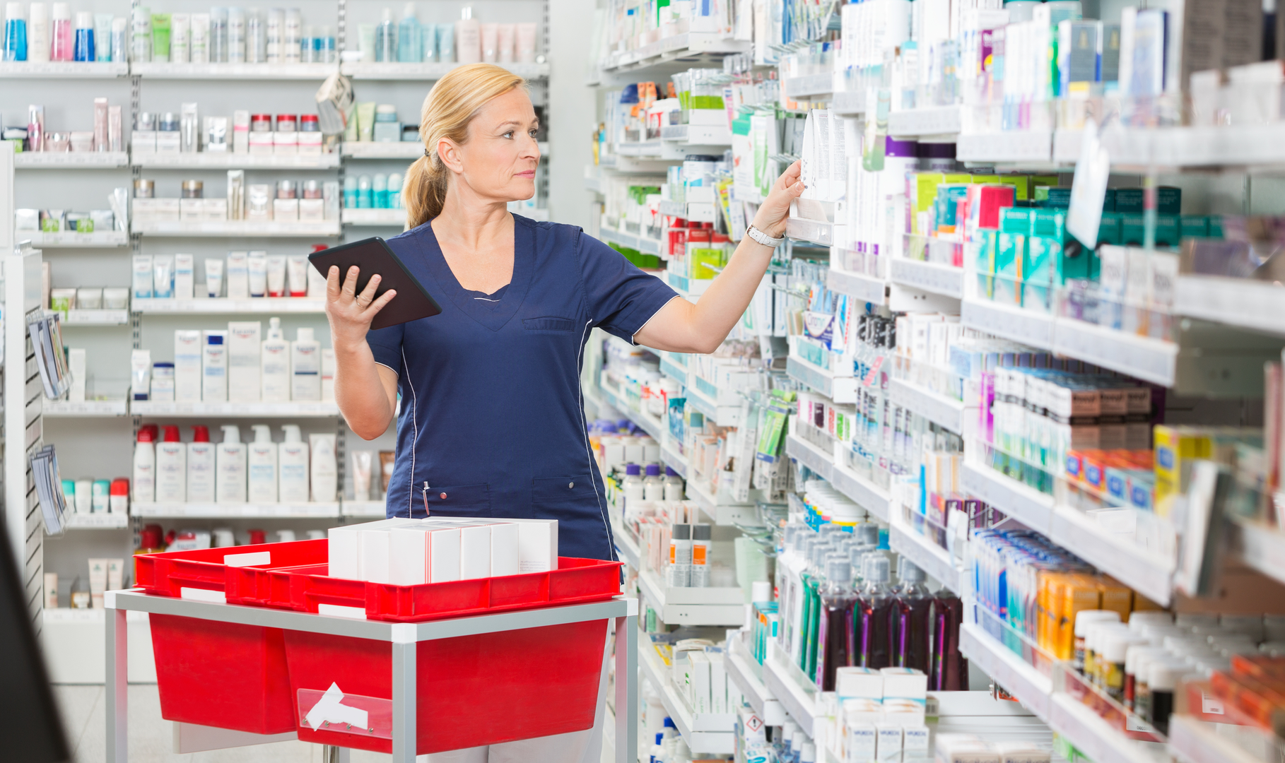 A pharmacy technician referencing data on a tablet and restocking a pharmacy's front-end shelves accordingly