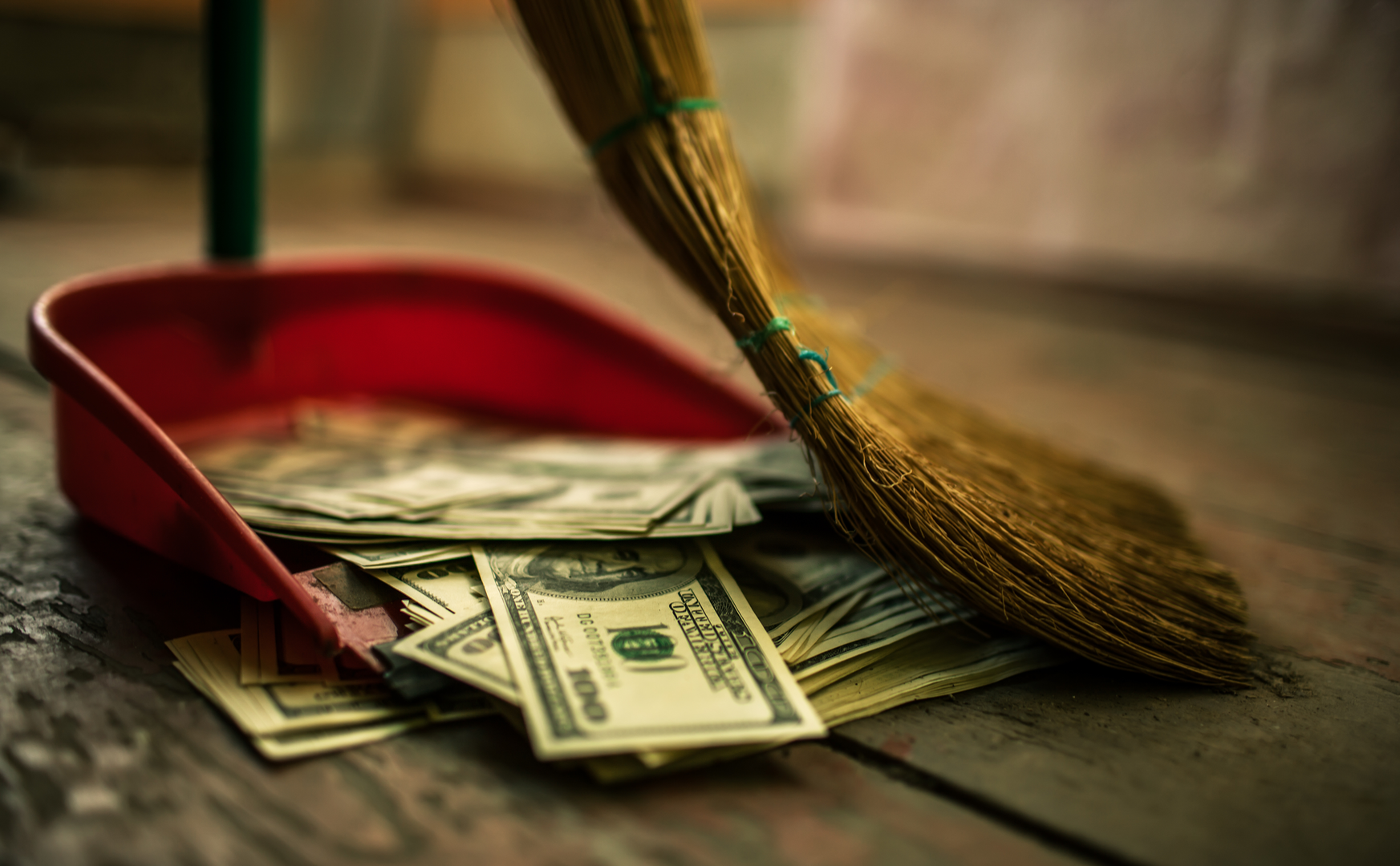 Someone using a broom to sweep dollar bills into a dust pan